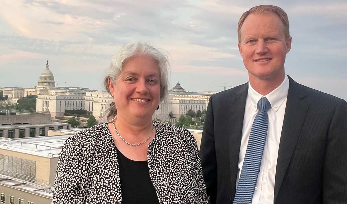 Joan Ramage and Jed Hancock pose for a photo on a rooftop in Washington, D.C. Behind them is the Capital building and the sun is setting.